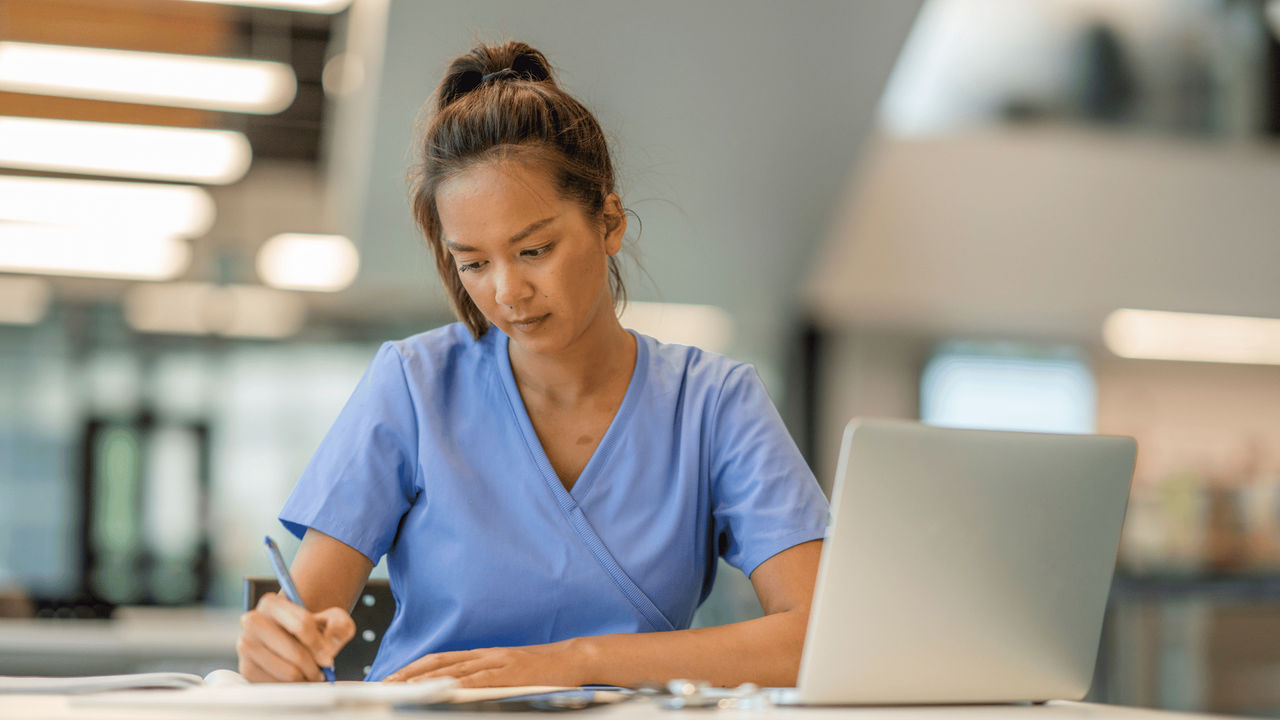 A UC Online nursing student, wearing scrubs, reviews her written study notes while working at a laptop.