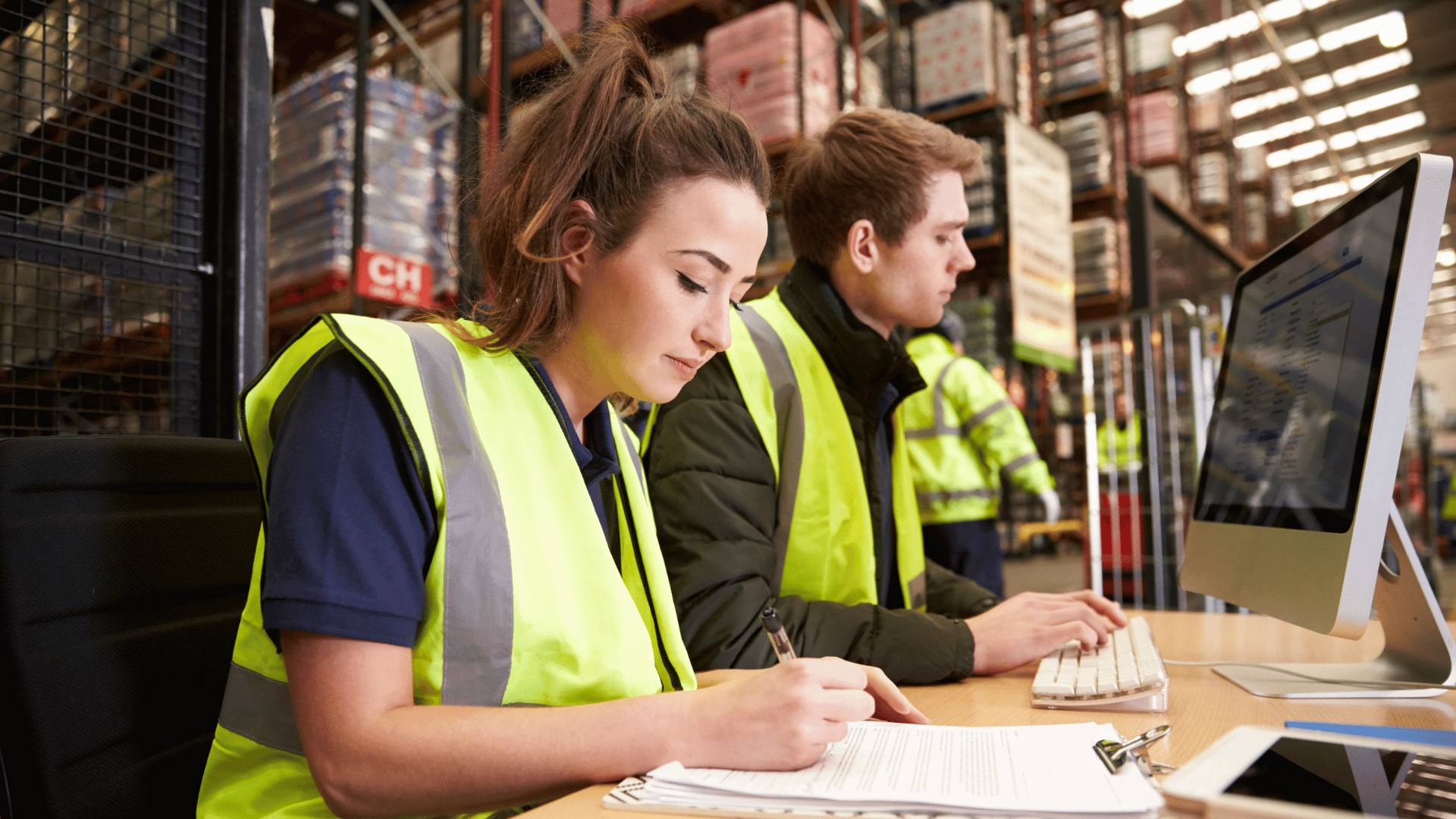 Two warehouse administration workers, wearing personal protective clothing, complete paperwork at a desk.