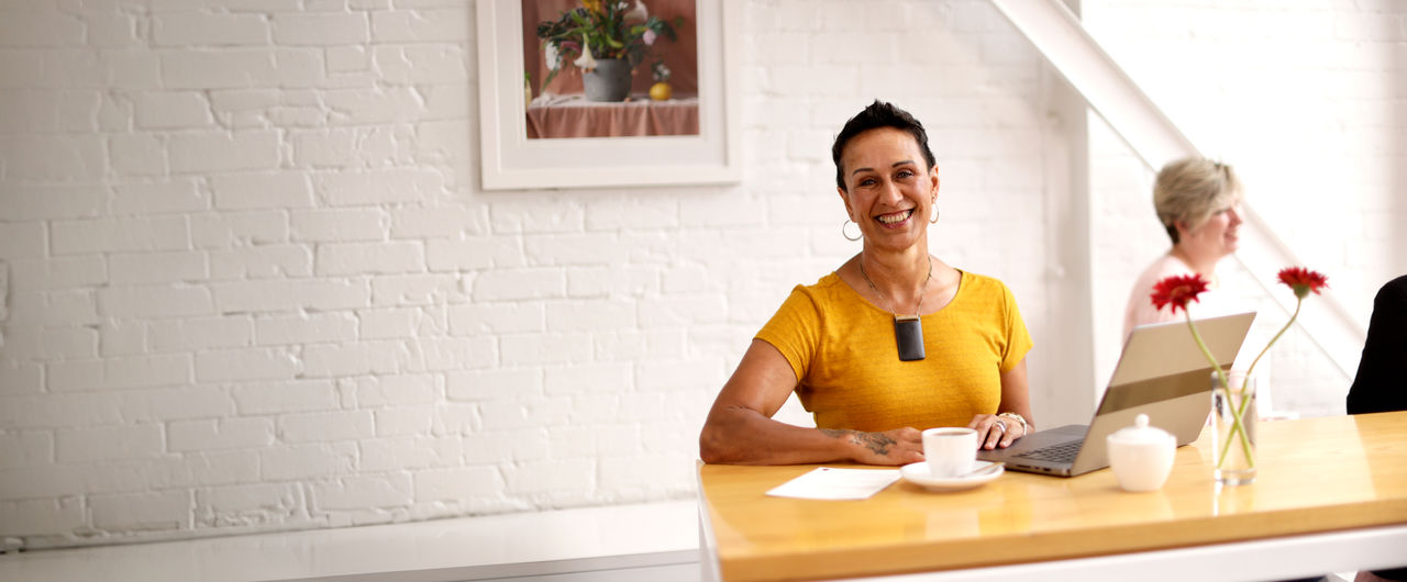 A UC Online student, working on a laptop in a light, bright cafe, smiles broadly at the camera.