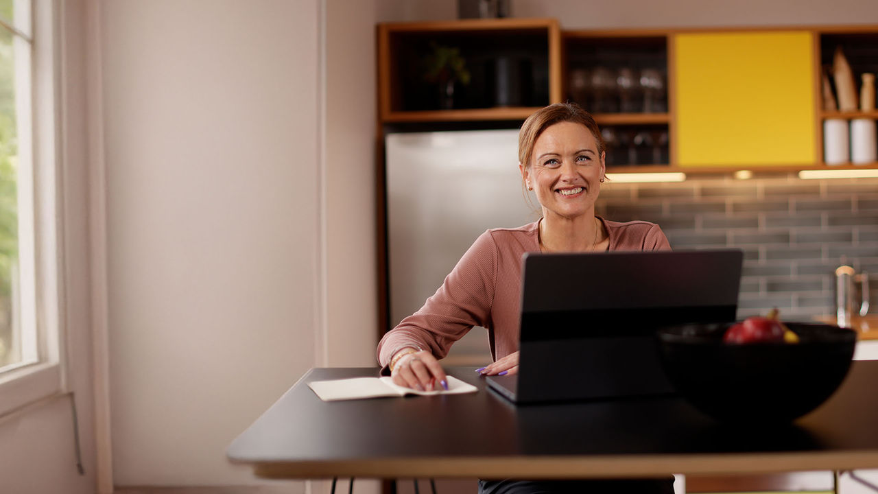 A UC Online student, working on a laptop in a light, bright cafe, smiles broadly at the camera.