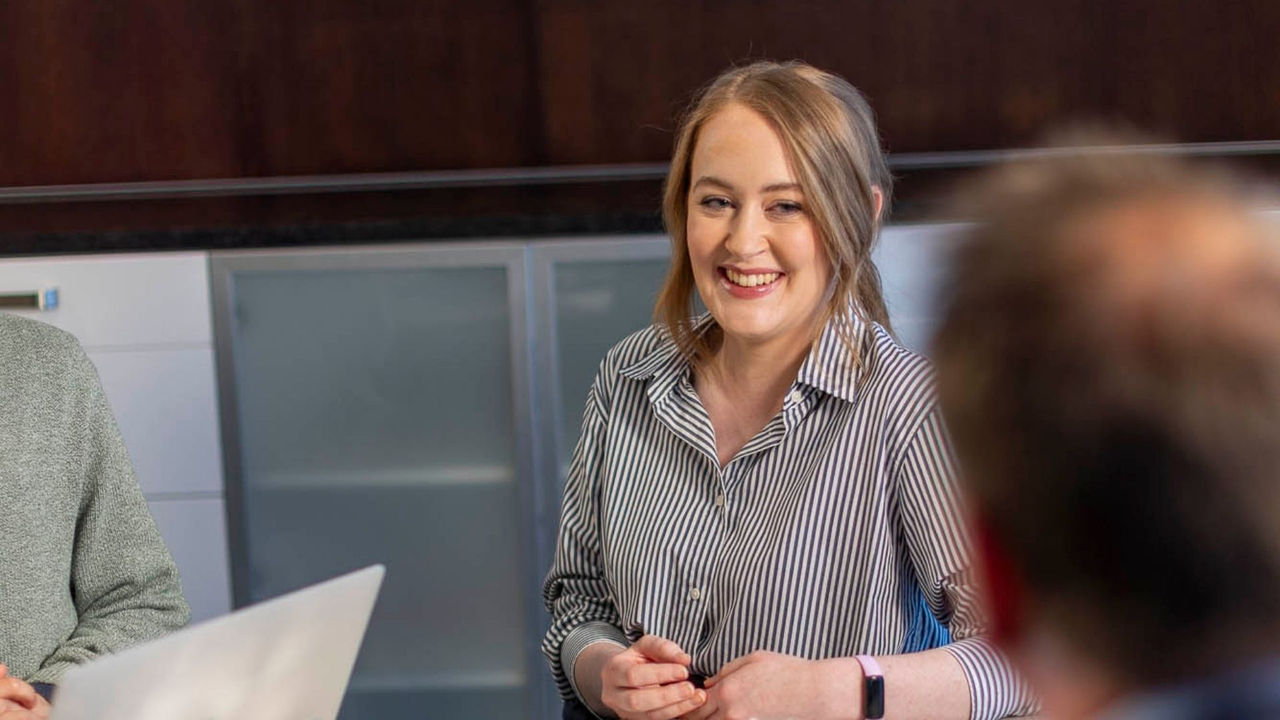 Woman in striped shirt talks to her coworkers in a boardroom about her coursework as part of her Master of AI. 