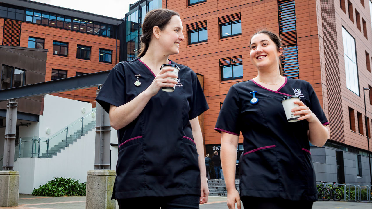 Two UC Online nursing students, dressed in scrubs, walk through the grounds of a hospital during a clinical placement.