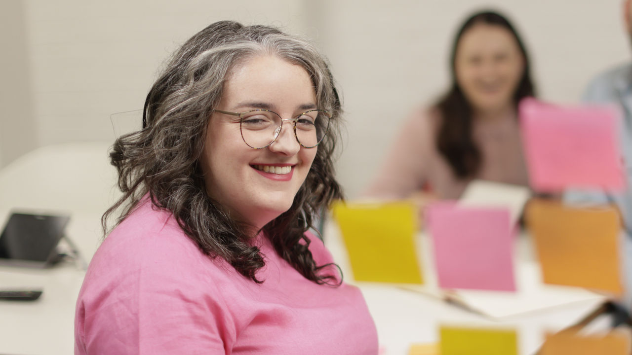  A smiling woman uses a board of coloured sticky notes to highlight data trends for her colleagues. 