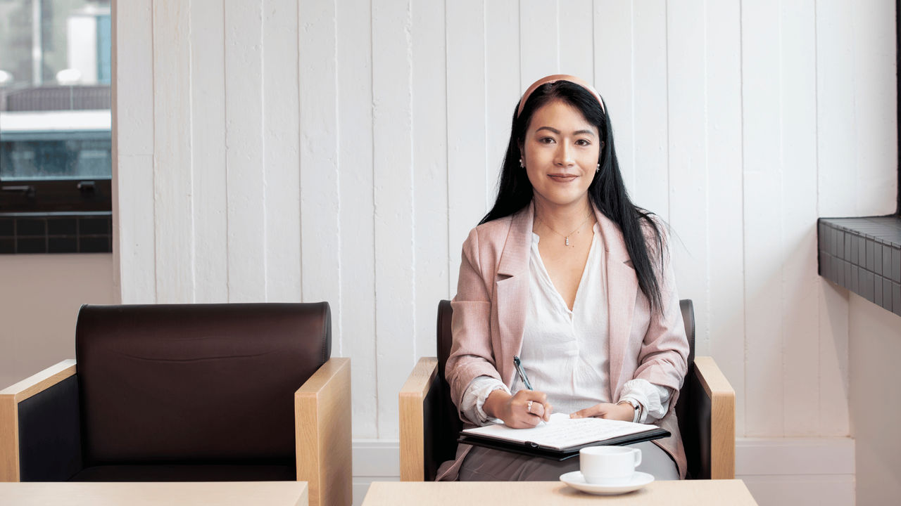 Woman sitting at a desk smiling into the camera while writing on a sheet of paper