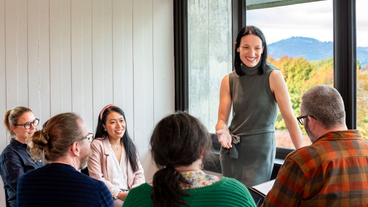 Woman sitting at a desk smiling into the camera while writing on a sheet of paper