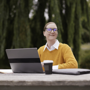 A UC Online Cyber Security student sits at a picnic table in a park, completing online study on her laptop.