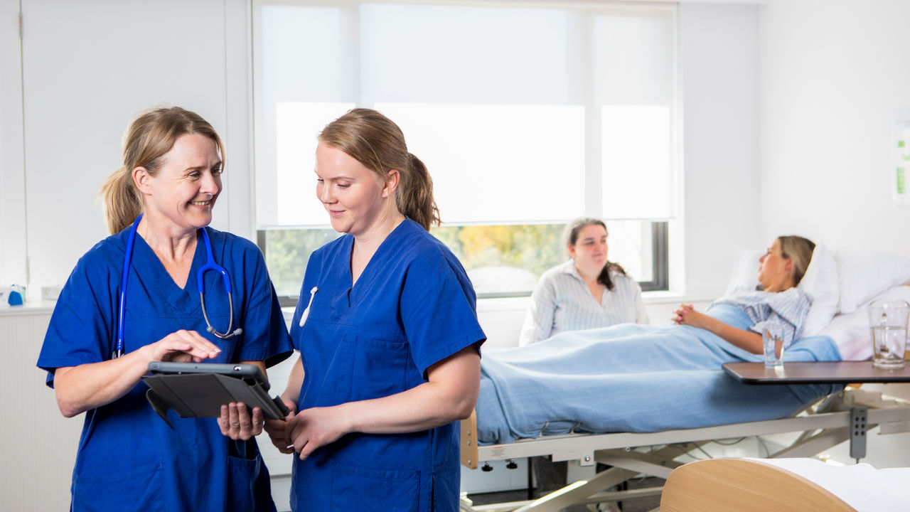 Two nurses talking while looking at an iPad, patient behind them with a friend