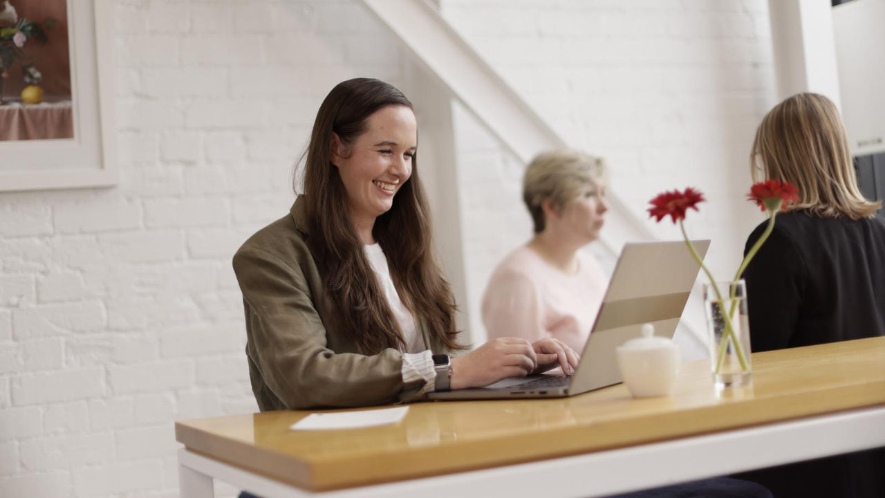 A smiling woman with long dark hair uses her laptop to study from her workplace, with colleagues visible in the background.